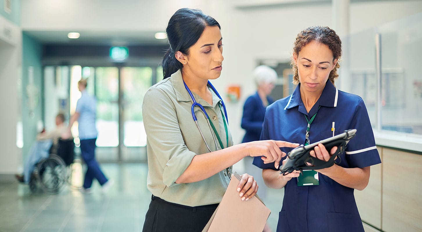Two healthcare professionals in scrubs reviewing information on a tablet in a hospital setting.